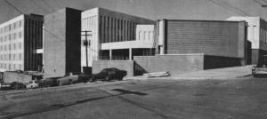 black and white photo of connected buildings of brick and concrete, with some construction details unfinished. Cars of the era are parked along the street.